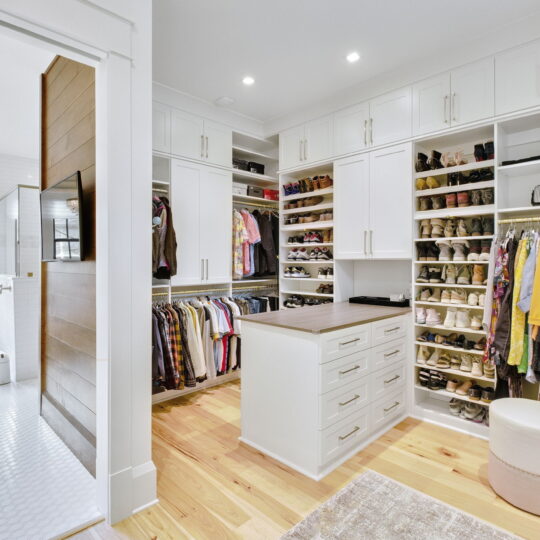 A spacious walk-in closet with white cabinetry filled with clothes, shoes, and accessories. A central island with drawers is surrounded by hanging garments, shelves, and closed cabinets. A glimpse of a bright bathroom is visible through an open doorway.