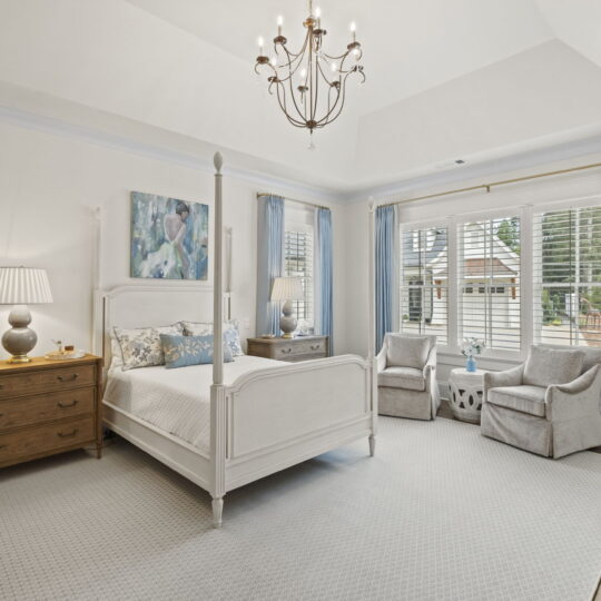 A spacious bedroom with a white bed, wooden nightstand, table lamp, and a large window with blue curtains. Two gray armchairs and a small table sit by the window. A chandelier hangs from the ceiling, illuminating the room.