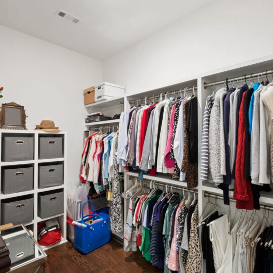 A walk-in closet with two racks of organized clothing, including shirts, blouses, and jackets. Shelves hold fabric bins and boxes. A blue storage bin and a mirror with a hat on top are also visible. The floor is hardwood.