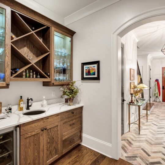 A home interior featuring a small bar area with wooden cabinets and glassware. To the right, an open hallway leads to a front door. A herringbone-patterned floor and pendant lights add elegance. A colorful artwork and coat rack are visible in the hallway.