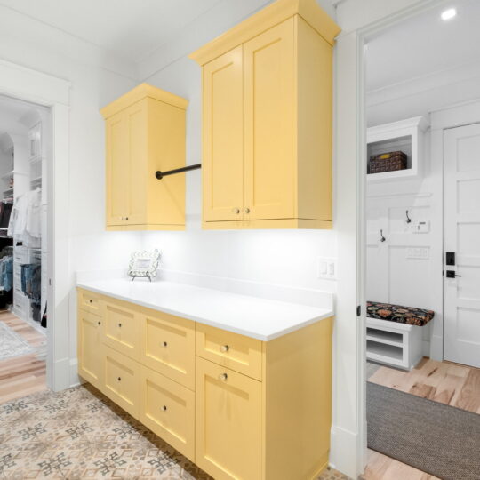 A bright laundry room features vibrant yellow cabinets with white countertops. On the left, theres a glimpse of a walk-in closet with clothes and a rug. The rooms floor is light wood, and theres a rug with a geometric pattern.