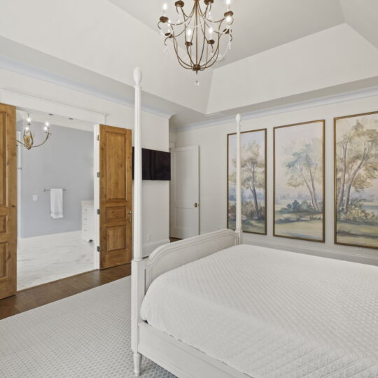 Elegant bedroom with a white four-poster bed, quilted white bedding, and pillows. Wooden double doors open to a bright en-suite bathroom. Three large tree-themed paintings and a chandelier add style. White and natural wood tones dominate the decor.