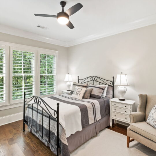 Cozy bedroom with a metal bed frame, layered with striped bedding and a decorative pillow. A beige armchair with pillows sits beside a white nightstand and lamp. Large windows feature plantation shutters, offering a view of lush greenery outside.