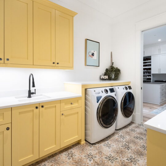 Laundry room with yellow cabinets, a white countertop, and a white sink with a black faucet. Two front-loading washers are side by side. The floor has patterned tiles, and a hallway leads to a room with shelves in the background.