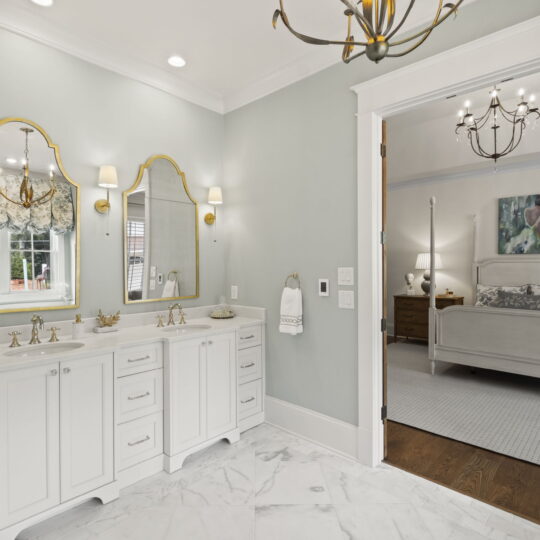 Elegant bathroom with white double vanity, gold-framed mirrors, and gold light fixtures. Marble floor leads to a bedroom featuring a white four-poster bed, chandelier, and artwork. The walls are light gray, and the room is well-lit by natural light.