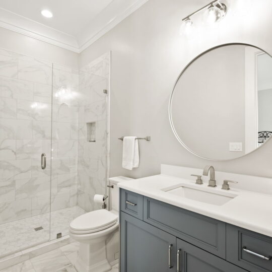 A modern bathroom with white marble tile flooring and walls. It features a glass-enclosed shower, a round mirror above a white sink with blue cabinetry, and a wall light fixture. A towel hangs beside the sink.