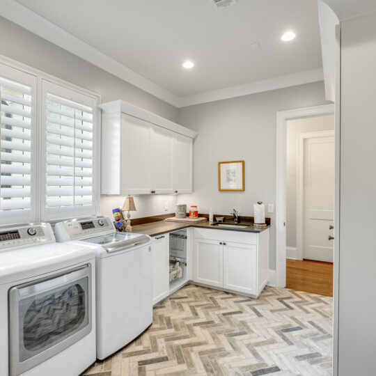 Modern laundry room with white cabinets and wooden countertops. Features a front-loading washing machine and dryer on a herringbone-patterned tile floor. A window with plantation shutters lets in natural light. A small sink area is visible.