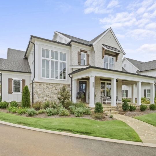 Modern two-story house with gray siding, large windows, and a covered porch. The home is surrounded by a landscaped garden with various shrubs and plants, and a paved walkway leads to the entrance. A car is parked on the driveway.