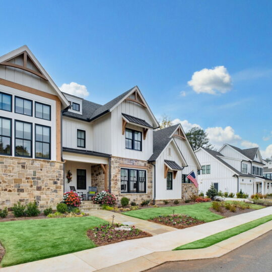 A suburban neighborhood with modern two-story houses. The homes have stone and siding exteriors, black-trimmed windows, and manicured lawns. An American flag hangs near one entrance. The sky is clear with a few clouds.