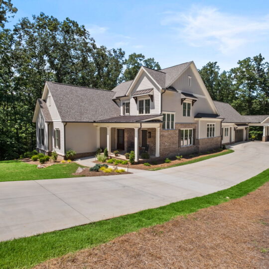 A large two-story house with a stone and beige siding exterior, surrounded by trees. It has a spacious driveway and well-maintained lawn, with a covered porch and dormer windows. The sky is clear and blue.