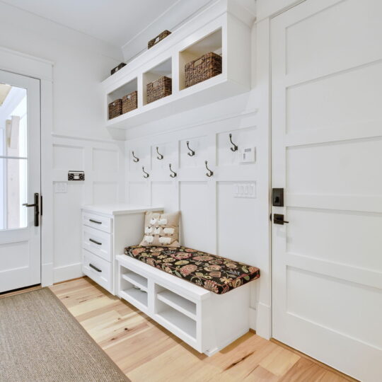 A bright mudroom with white walls and wooden floors, featuring a cushioned bench, storage cubbies above, and multiple hooks for hanging items. A glass door and a large mat are visible, along with a basket of flowers outside.