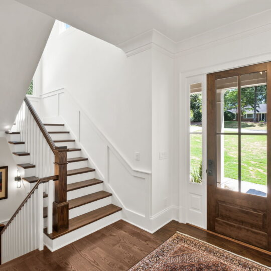 A bright, spacious entryway features a wooden staircase with white railings. A large wooden front door with glass panels opens to a sunlit yard. A framed picture hangs on the wall, and a patterned rug is on the wooden floor.