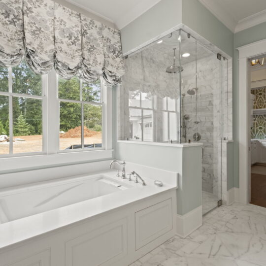 Luxurious bathroom with a marble floor, large bathtub, and glass-enclosed shower. Floral-patterned curtains adorn the window, showcasing a view of trees and a dirt pile outside. A doorway leads to a hallway with patterned wallpaper.