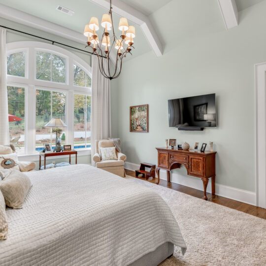 A spacious bedroom featuring a large bed with a white quilt and patterned pillows, a TV mounted on the wall, and a wooden desk. There are tall windows with curtains and a door leading to an outdoor patio area visible in the background.