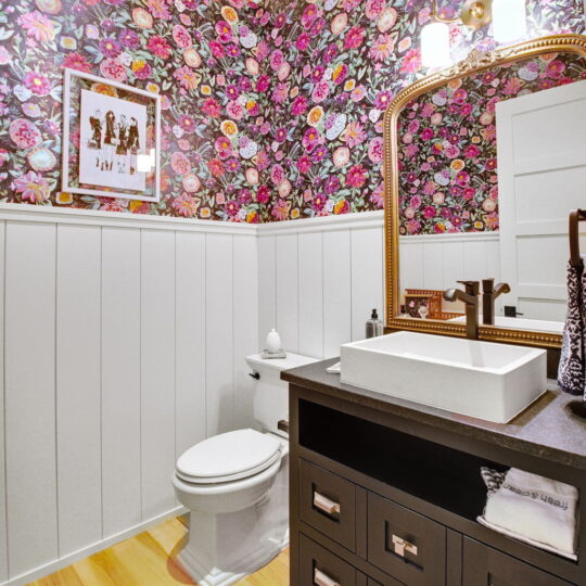 Bathroom with floral wallpaper, a white wainscoting on lower walls, and a wooden vanity with a square sink. Theres a framed artwork above the toilet. The floor is light wood, and two wall lights are above a large mirror.