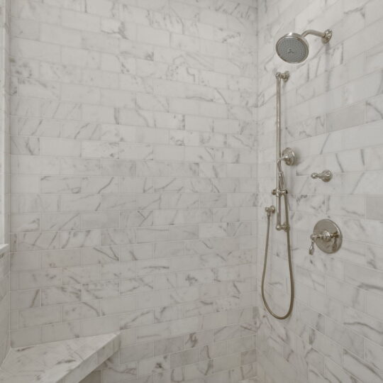 A marble-tiled shower with a large round overhead showerhead and a handheld shower on the wall. It features a built-in corner bench and hexagonal floor tiles. The walls have a white and gray marble pattern.