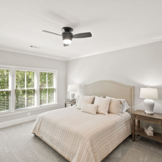 A bright bedroom with a large bed featuring a light beige headboard and matching bedding. Two wooden nightstands flank the bed, each with a lamp. A ceiling fan is above, and a window with blinds offers a view of greenery outside.