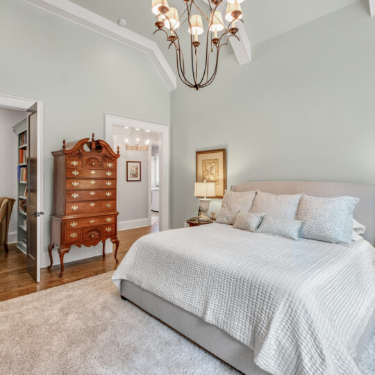 A cozy bedroom with a large gray bed featuring multiple pillows. A chandelier hangs from the ceiling. Theres a wooden dresser, a painting on the wall, and a nightstand with a lamp. An open door reveals a small seating area with a striped armchair.