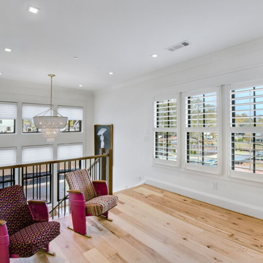 Two patterned armchairs with pink bases sit on a wooden floor in a bright room. Large windows with shutters let in natural light. A modern chandelier hangs above the seating area, and a wooden railing overlooks a lower floor.