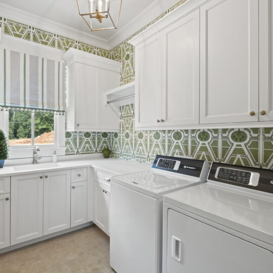 A bright laundry room with white cabinets and appliances. The walls feature green geometric wallpaper. A window with a striped curtain lets in natural light. A small potted plant sits on the countertop.