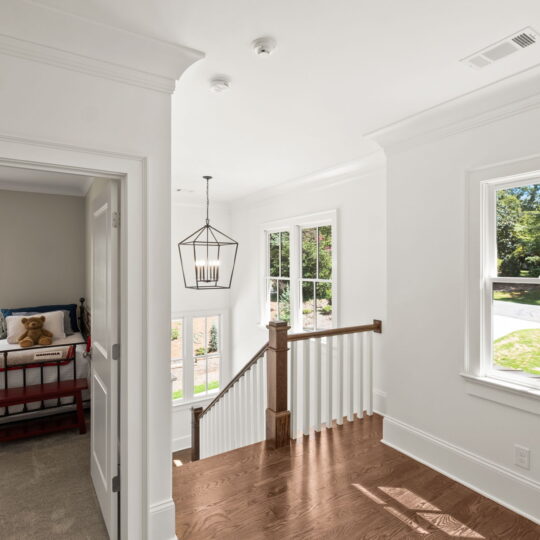 A bright, sunlit hallway with wooden floors and white walls. It leads to a child’s bedroom with a red bed, teddy bear, and a side table with a lamp. A staircase with white railing and an elegant geometric pendant light is also visible.