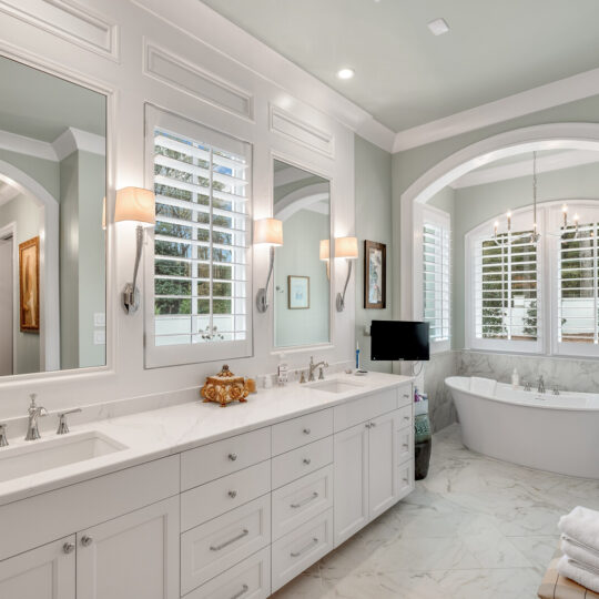 Luxurious bathroom with freestanding bathtub, double vanity with mirrors, two wall-mounted lights, shutters on windows, and marble floor. Neutral tones and elegant decor create a serene atmosphere.