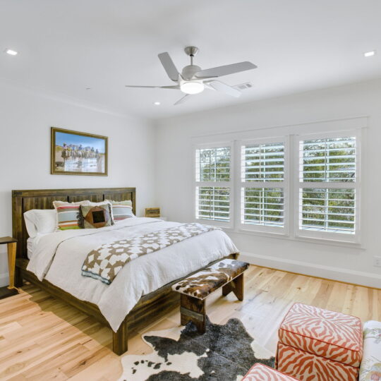 A bright bedroom with a large bed featuring a geometric-patterned blanket. Theres a ceiling fan, windows with shutters, a framed art piece above the bed, a small bench, and a cozy chair with an orange patterned ottoman. Light wooden floor.