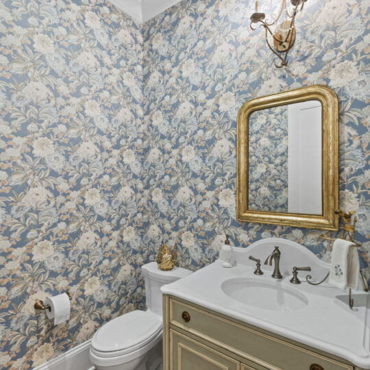 A small bathroom with floral wallpaper, featuring a white toilet, gold-framed mirror, and cream-colored vanity with a white countertop. A gold pineapple decor sits on the tank, with dual wall-mounted lights above the mirror. Hardwood floor adds warmth.