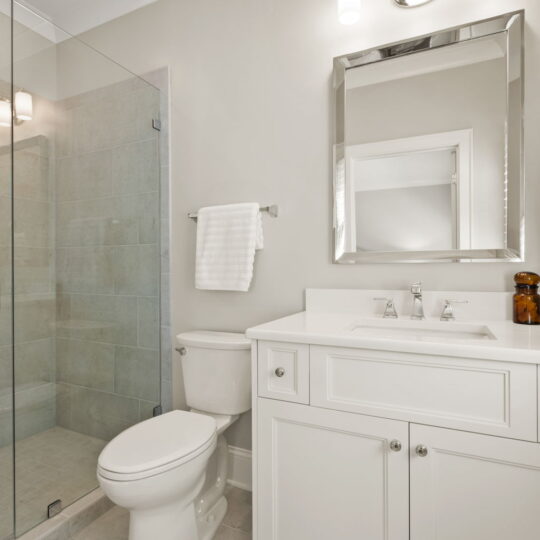 A modern bathroom with a glass-enclosed shower, white toilet, and a white sink vanity with a square mirror above. A towel hangs next to the toilet, and two lights are mounted above the mirror. The floor is tiled in light gray.