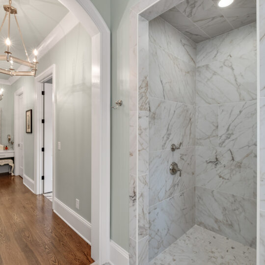 A hallway features hardwood floors, light green walls, and a chandelier. There is a marble tiled shower on the right, and a dressing table with a mirror and drawers at the end of the hall.