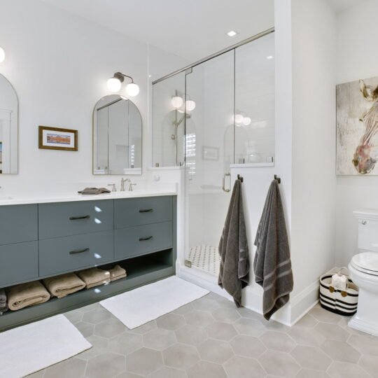 Modern bathroom with a double-sink vanity, blue cabinets, and towels neatly stored underneath. A large mirror with two globe lights above each sink. Glass-enclosed shower with white tiles, a toilet, and zebra artwork on the wall. Light gray hexagonal floor tiles.