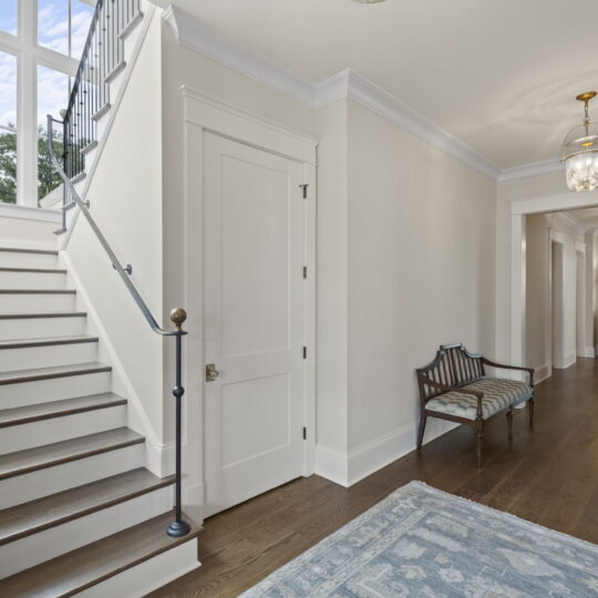 A bright hallway with wooden floors, a staircase on the left, and a large window above. A wooden bench sits against the wall, and a rug covers part of the floor. There are elegant light fixtures on the ceiling.