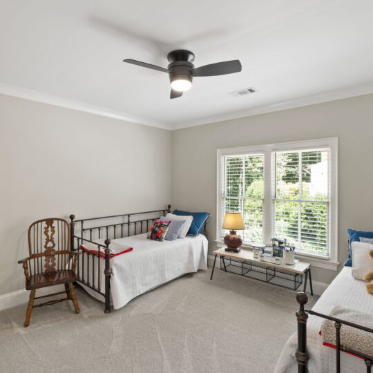 A bedroom with two metal-framed single beds, each with white bedding. A wooden chair is beside one bed. A window with white blinds lets in natural light. A small table with a lamp and books is placed between the beds. A ceiling fan is above.