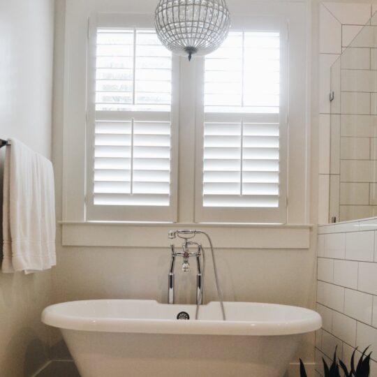 A bathroom with a clawfoot bathtub beneath shuttered windows. A spherical pendant light hangs from the ceiling. A white towel is on the left towel rack, and a small potted plant is on the floor near the tub.