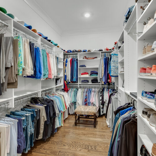 A well-organized walk-in closet with neatly arranged clothes on hangers, including shirts, pants, and jackets. Shelving on both sides displays folded clothes, hats, and shoes. A cushioned stool is in the center, and the floor is wooden.