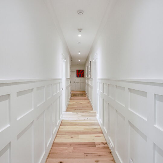 A long hallway with white paneled walls and wooden flooring, lit by ceiling lights. A red abstract painting hangs at the end of the hallway near a closed door. The perspective emphasizes the length and symmetry of the corridor.