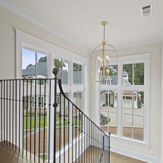 A staircase with wooden steps and black metal railing descends in a bright hallway with large windows. Outside, a paved area and greenery are visible. A chandelier hangs from the ceiling, adding elegance to the space.