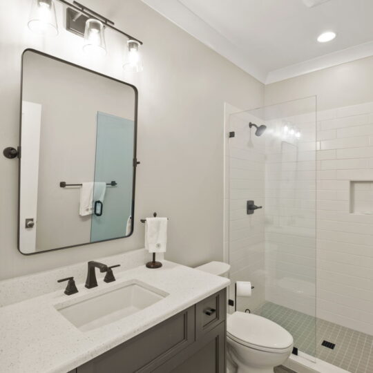 Modern bathroom with a white countertop, dark vanity, and black fixtures. A large mirror with three lights above it is on the wall. Theres a white toilet and a glass-enclosed shower with white subway tiles. A beige rug is on the wooden floor.