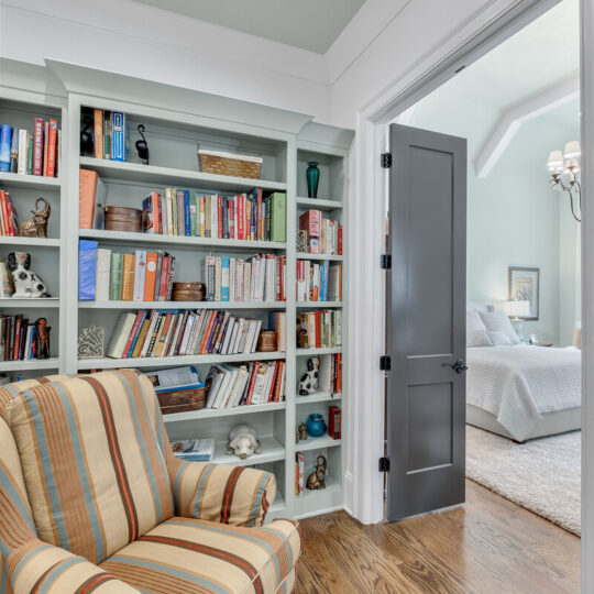 Cozy reading nook with a striped armchair in front of a filled bookshelf. The space leads to a bedroom visible through a partially open gray door, featuring a bed and elegant chandelier. The hardwood floor complements the soothing pastel wall colors.