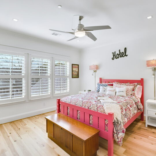 A bright bedroom featuring a pink bed with a colorful quilt, two decorative lamps, and a wooden bench at the foot. A window with white shutters lets in natural light. The word Hotel is displayed above the bed. The floor is light wood.