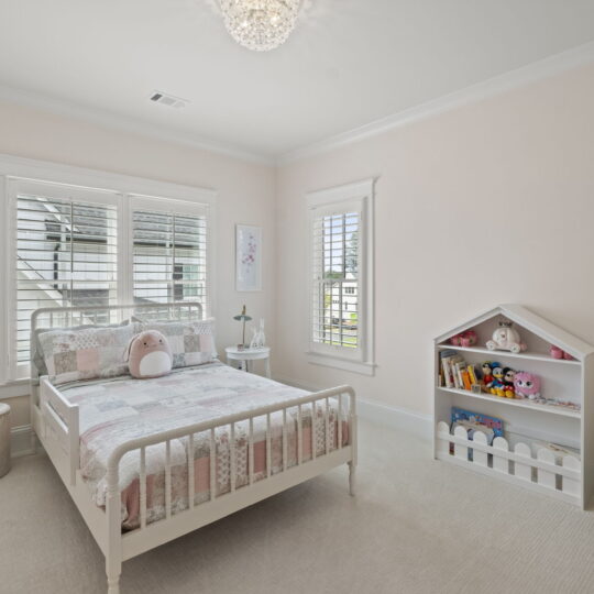 A cozy childrens bedroom with a white bed featuring a patchwork quilt and plush toys. There are two small framed pictures on the walls, a white bookshelf with toys, and a green armchair. Soft natural light filters through the windows with white shutters.