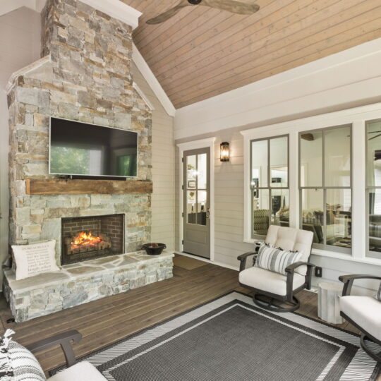 A cozy outdoor living space featuring a stone fireplace with a TV mounted above. Several cushioned chairs surround the area, accented with striped pillows. A small decorated Christmas tree stands to the side. Large windows and a ceiling fan complete the scene.