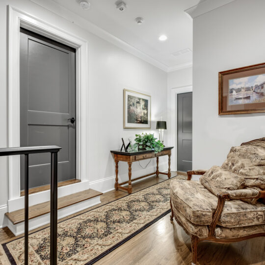 A traditional hallway with a patterned rug on hardwood floors, a plush armchair, and a wooden side table adorned with a plant and lamp. Two doors are visible, and framed paintings are hung on the light-colored walls.