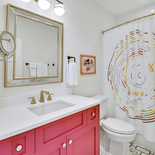 A modern bathroom with a bright pink vanity with a white countertop. Above it is a rectangular mirror with lights. The shower curtain features a colorful, circular pattern. White towels hang on the wall, and the floor has a geometric tile design.