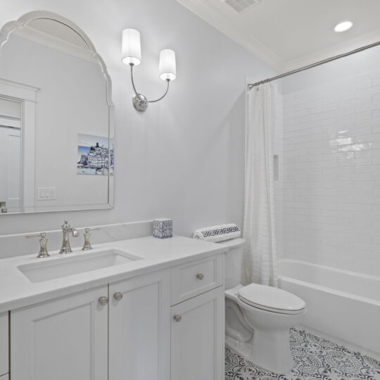 A bright, modern bathroom with white cabinetry, a large mirror, dual wall sconces, and a tiled bathtub with curtain. The floor has a patterned tile design, and a framed picture hangs on the wall.