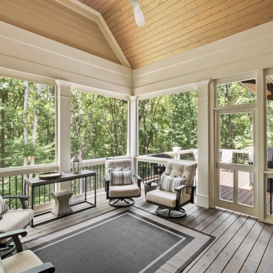 A cozy screened-in porch features cushioned chairs, a small table, and a patterned rug. The porch overlooks a lush, green forest, providing a serene and natural backdrop. The space has a wooden ceiling and a modern, airy design.