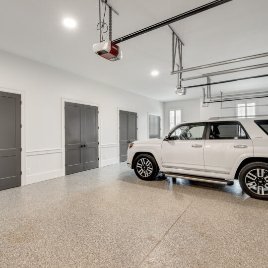 Spacious garage interior with a white SUV parked. The garage features a polished, speckled floor, white walls, and multiple dark gray doors. Ceiling-mounted garage door openers are visible, and natural light enters through windows and open doors.