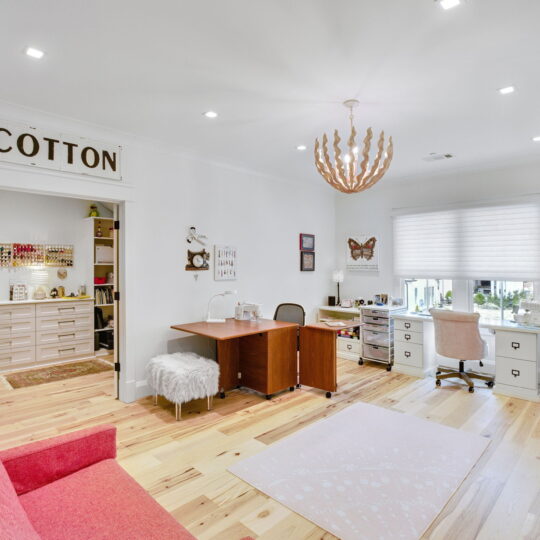 A bright craft room with a red sofa, a wooden desk with chairs, and a white storage cabinet. Shelves are filled with thread spools. A modern chandelier hangs from the ceiling, and large windows provide natural light. A COTTON sign is above the door.