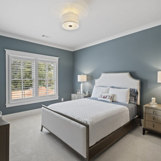 A modern bedroom with teal walls, a white bed with beige headboard, and a pair of nightstands with lamps. A dresser with flowers and a TV are on the left. Natural light streams through a window with blinds.