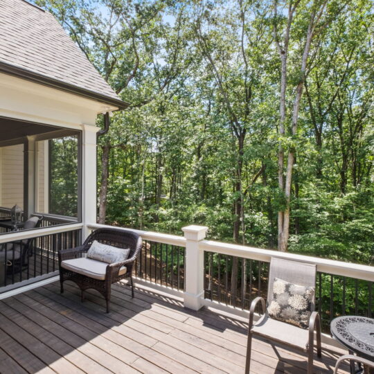 A wooden deck overlooks a lush forest. The deck features a woven chair with a cushion, a round metal table with two matching chairs, and a railing with a towel draped over it. The adjacent house has a screened-in porch.